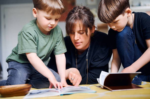 Молода біла мати проводить час вдома з синами і читає книги на підлозі. Щасливий батько грає з дітьми дошкільного віку. Концепція домашньої освіти
 - Фото, зображення