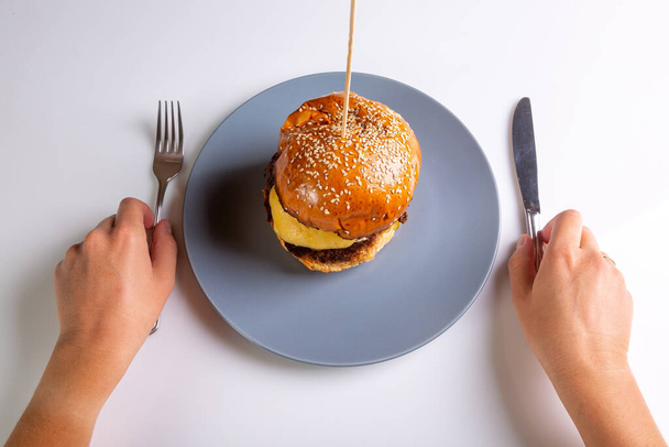 Hausgemachter Cheeseburger auf einem grauen Teller und eine Person mit Besteck in der Hand auf weißem Hintergrund. Hände im Rahmen. Blick von oben. Horizontale Ausrichtung. Hochwertiges Foto - Foto, Bild