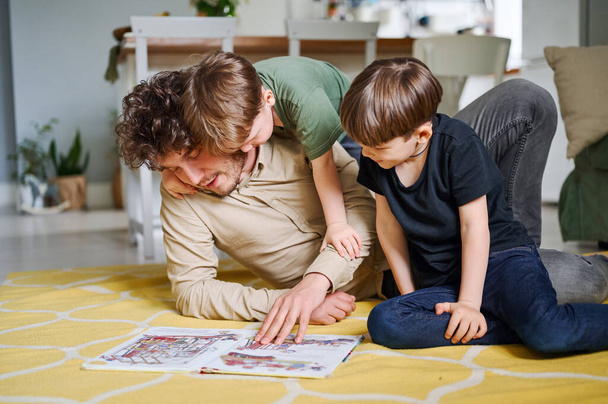 二人の息子を持つ若い父読書本すぐに床、幸せな親は子供と遊んで一緒に時間を過ごす - 写真・画像