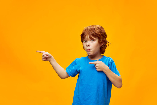 red-haired boy in a blue t-shirt shows his index fingers to the side funny facial expression  - Fotografie, Obrázek