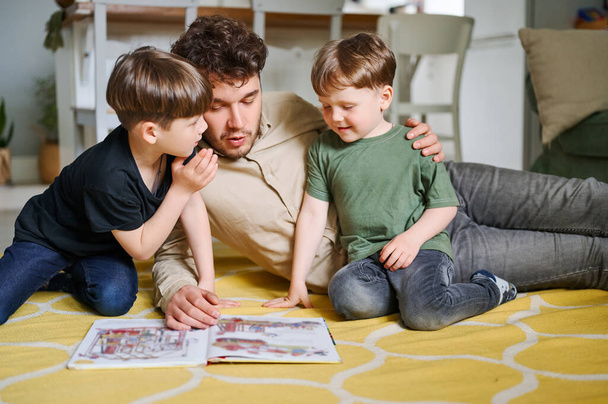 Vater mit Söhnen liest Buch, verbringt Zeit zu Hause und liegt auf dem Boden - Foto, Bild