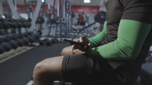 Onbekende zwarte man typt snel bericht in de telefoon in de sportschool. Sportfaciliteiten op de achtergrond. Smartphone op handen. - Video