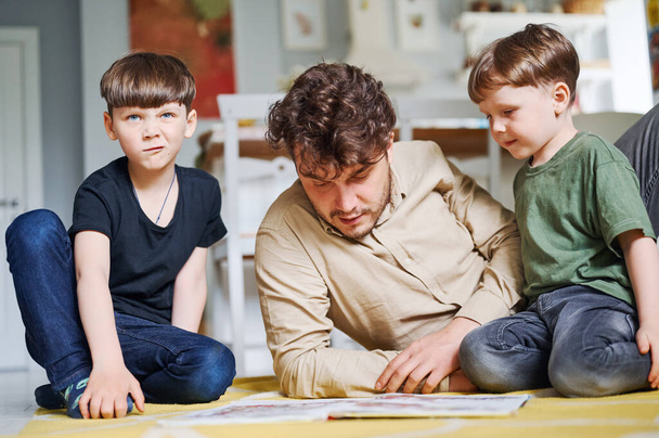 Vater mit Söhnen liest Buch, verbringt Zeit zu Hause und liegt auf dem Boden - Foto, Bild