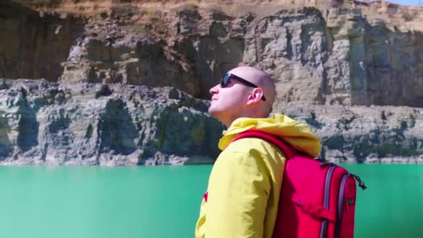 Reiziger onderzoekt het gebied. Zicht van achteren. Stijlvolle kerel met een rode rugzak gekleed in een gele regenjas staat tegen de achtergrond van een blauw meer in een stenen canyon. - Video