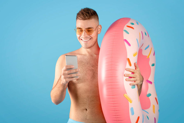Online tourist agency. Young man with inflatable ring and smartphone booking tropical tour on web, blue background - Фото, изображение