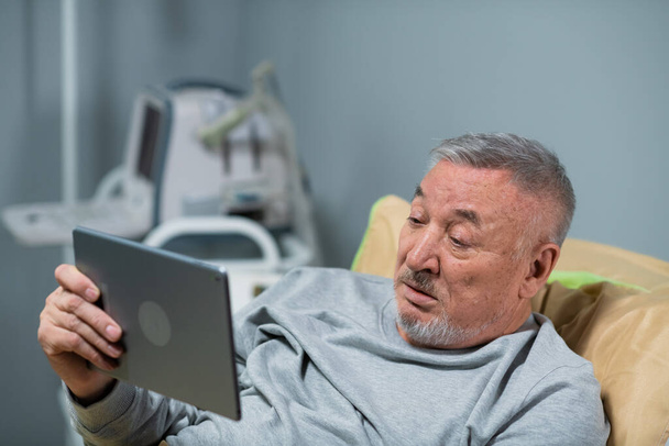 Starzec na oddziale szpitalnym ogląda mecze sportowe za pomocą tabletu. - Zdjęcie, obraz