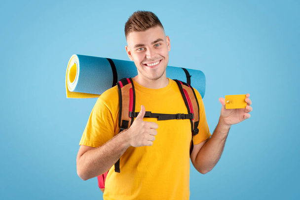 Travel budget. Cool millennial guy with backpack and credit card showing thumb up gesture over blue studio background - Photo, Image
