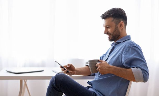 Рабочий перерыв. Расслабленный мужчина фрилансер с помощью смартфона и питьевой кофе дома - Фото, изображение