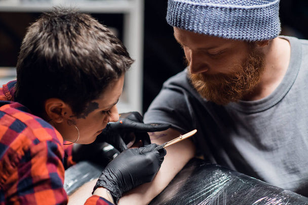 A tattoo artist with short hair and piercings is preparing to get a tattoo on a mans arm in a tattoo parlor - Photo, Image
