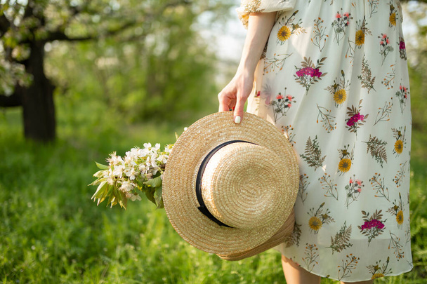 A girl in a light summer sundress walks in a spring green park enjoying the blooming nature, carries a wicker hat and flowers in her hand. - Foto, afbeelding