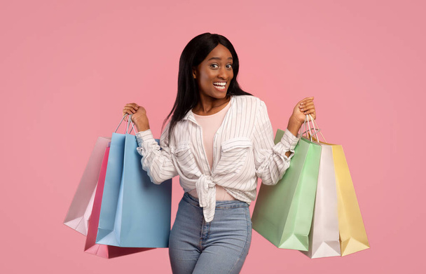 Концепция большой продажи. Счастливая черная молодая женщина в повседневной одежде держит сумки с покупками на розовом фоне студии - Фото, изображение