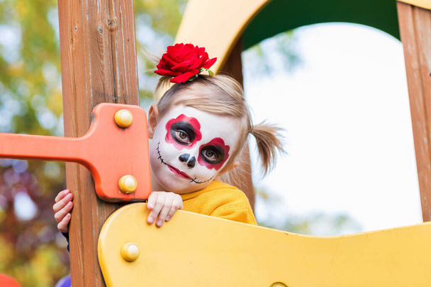 Маленька дошкільна дівчинка з намальованим обличчям піднялася на драбину на ігровому майданчику, відзначаючи Хелловін або Мексиканський день мертвих.. - Фото, зображення