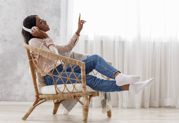 Mujer africana alegre cantando mientras escucha música con auriculares inalámbricos en casa - Foto, imagen