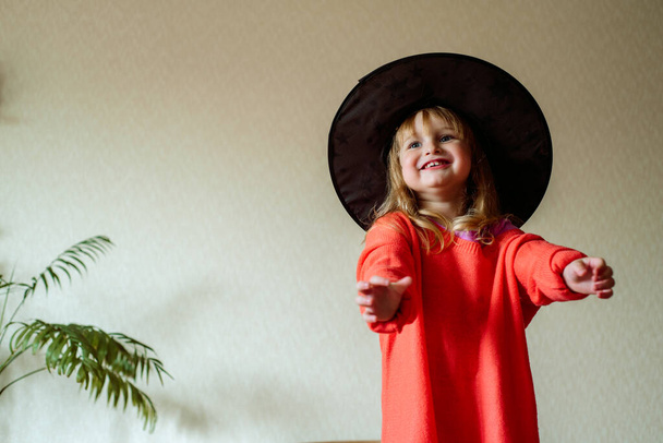 little girl in a witch hat with an orange ball in her hand on a yellow background. Halloween concept.Active games at home. Laughs emotionally and jumps on the bed. - Foto, imagen