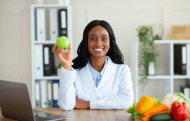 Dietista femminile nera positiva che tiene frutta di mela e guarda la fotocamera in clinica - Foto, immagini