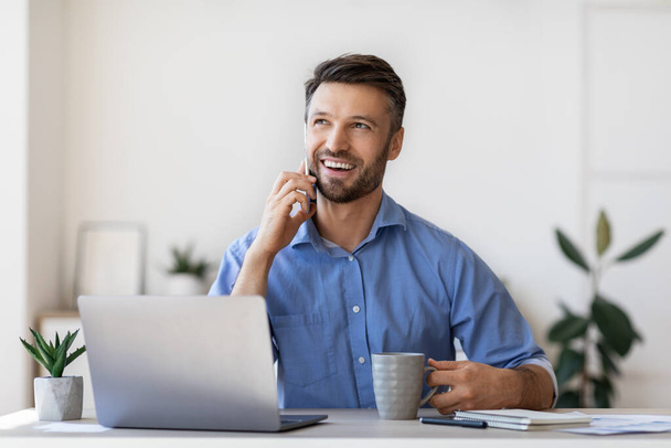 Веселый мужчина-предприниматель разговаривает по мобильному телефону и пьет кофе в офисе - Фото, изображение