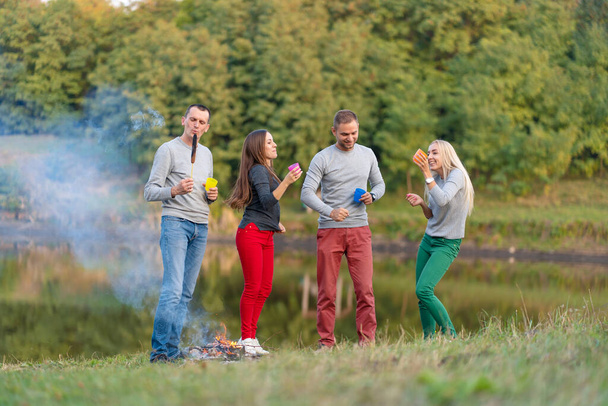 Picnic with friends in at lake near bonfire. Company friends having hike picnic nature background. Hikers relaxing during drink time. Summer picnic. Fun time with friends. - 写真・画像