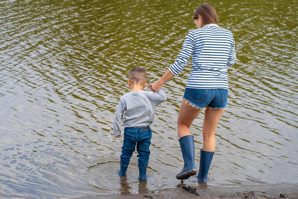 Mom with a small son walks along the sandy shore of the lake in rubber boots. Hanging out with children in nature, away from the city. - Fotografie, Obrázek