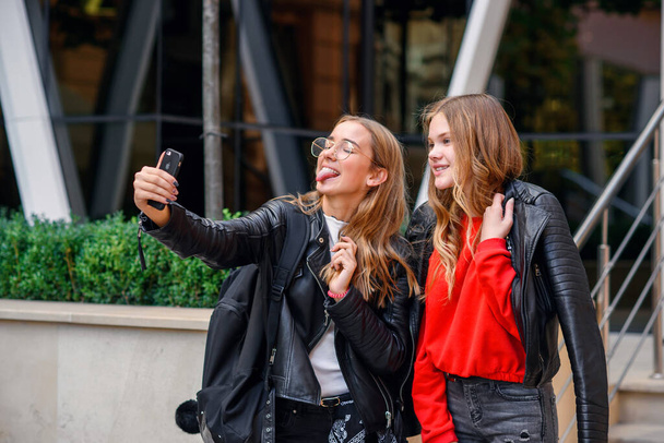 Two stylish happy teenage girls with smartphone making selfie near stylish modern building on the street. - Fotoğraf, Görsel