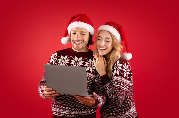 Мила дівчина та її бойфренд в зимовому одязі для різдвяних подарунків онлайн на ноутбуку, червоне тло - Фото, зображення