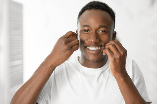 Ευτυχισμένος Αφροαμερικανός άνθρωπος Flossing δόντια στέκεται στο σύγχρονο μπάνιο - Φωτογραφία, εικόνα