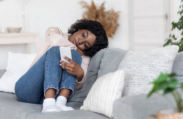 Osamělost. Depressed černošky dáma sedí na gauči s chytrým telefonem, Loking na obrazovce - Fotografie, Obrázek