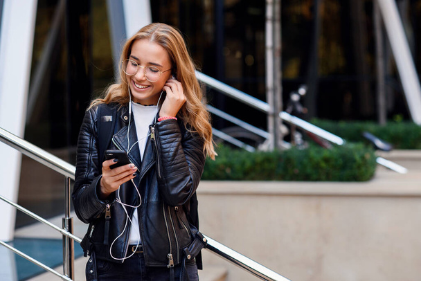 Young happy beautiful girl in urban background listens to music with headphones. Joyful girl walks on the street. - Photo, Image