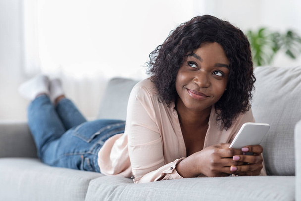 Mujer africana pensativa usando teléfono inteligente mientras está acostado en el cómodo sofá en casa - Foto, Imagen
