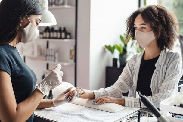 New normal and fight against virus in beauty salon. Asian female master in protective mask works with nails and disinfect hands with antiseptic of african american lady - Photo, Image