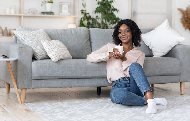 Ονειρεμένη Αφρο-Αμερικανίδα που χαλαρώνει στο σπίτι με ένα φλιτζάνι καφέ - Φωτογραφία, εικόνα