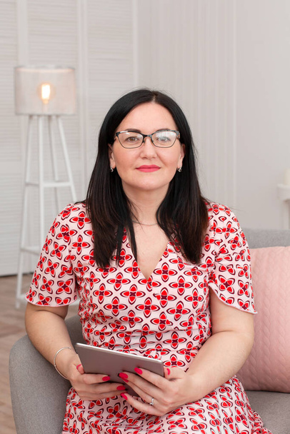 Portrait of a brunette woman in a red and white checkered dress, and glasses sits in a comfortable chair with an electronic tablet. - Foto, imagen