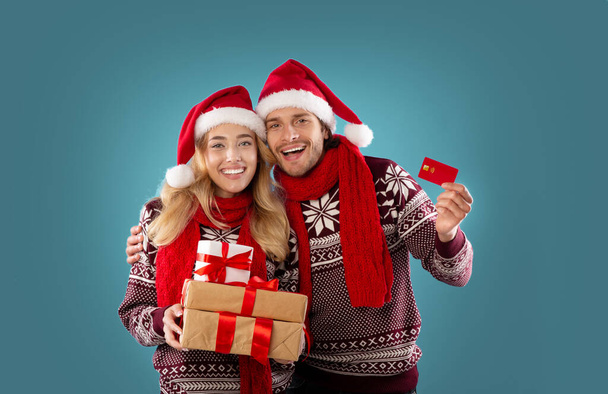 Christmas shopping. Young man and his girlfriend with pile of gifts and credit card on blue background - Photo, Image