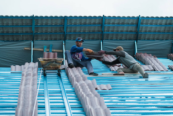 マレーシアのムアドザムシャー- 2020年10月3日:バケツとロープを使ってリフト屋根タイルを動かす2人の労働者が建物の屋根を設置する準備をする - 写真・画像