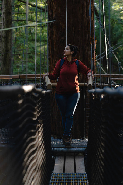 Nuori brunette nainen seisoo keskellä riippusilta on punainen puu metsä. Pystyvalokuvaus - Valokuva, kuva