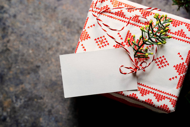 Χριστουγεννιάτικο κουτί δώρου τυλιγμένο σε εορταστικό χαρτί, με άδεια ετικέτα και κόκκινη ταινία, περιτριγυρισμένο από κλαδιά πεύκου σε μπλε φόντο grunge. Από ψηλά. Χριστουγεννιάτικη ιδέα στο σπίτι - Φωτογραφία, εικόνα
