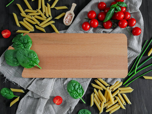 IItaliaanse macaroni penne met tomaten en kruiden ligt rond een snijplank op een zwarte achtergrond met een plaats voor tekst, close-up. - Foto, afbeelding