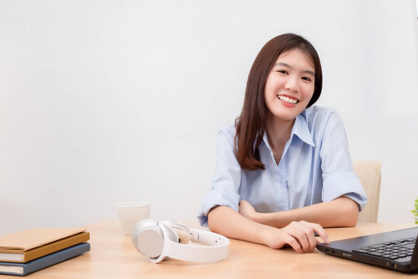 Mulher asiática bonita sorrindo no escritório de mesa com laptop, fones de ouvido, bloco de notas, caneca de café quente na mesa. - Foto, Imagem