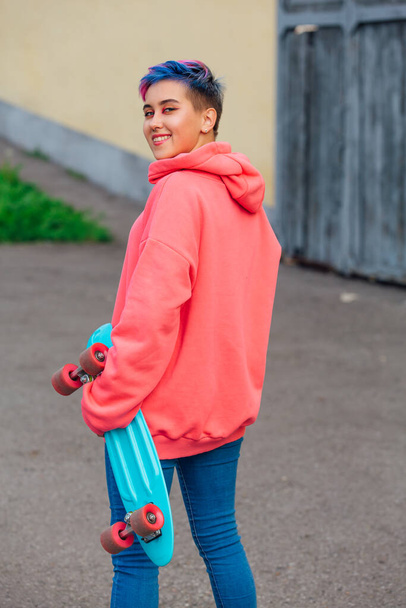 Stylish young woman with short colored hair holding blue plastic skateboard in hand. Youth concept. - Foto, imagen