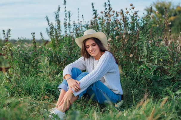 Young beautiful brunette woman dressed in a white sweater, jeans and cowboy straw hat sitting on green grass, smiling and laughing during sunset - Photo, Image