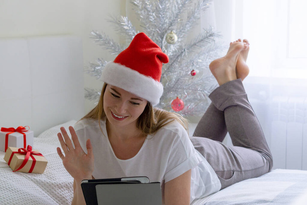 Noel çevrimiçi tebrikler. Noel Baba şapkalı neşeli kız yatakta uzanıyor ve tablet kullanarak video görüşmesi yapıyor. - Fotoğraf, Görsel