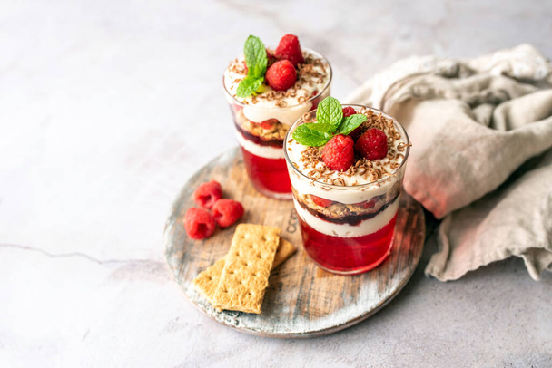 Dessert in a glass cup, with Jello covered in yogurt and topped with fresh raspberries. Mini dessert on light concrete background. Fruit parfait with jello and jelly. - Фото, зображення