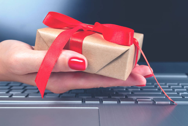 Womans hand holding present box with red bow and laptoptop computer as background behind it for your project. Seasonal sale, black friday and cyber monday concept. Online shopping - Fotografie, Obrázek