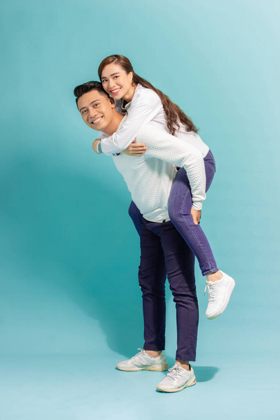 Full length portrait of a cheerful man carrying girlfriend on his back and having fun over blue wall - Foto, Bild