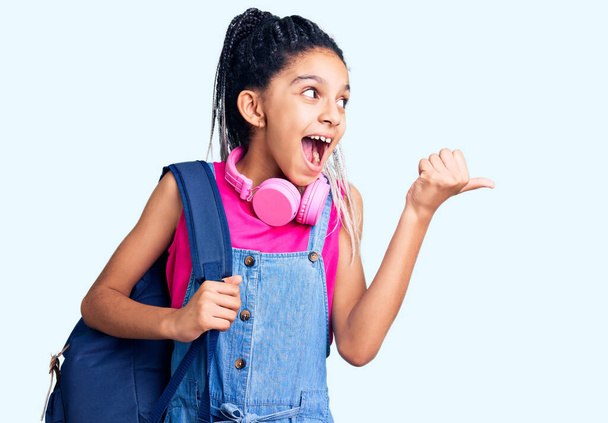 Menina afro-americana bonito segurando mochila estudantil usando mochila apontando polegar para o lado sorrindo feliz com a boca aberta  - Foto, Imagem