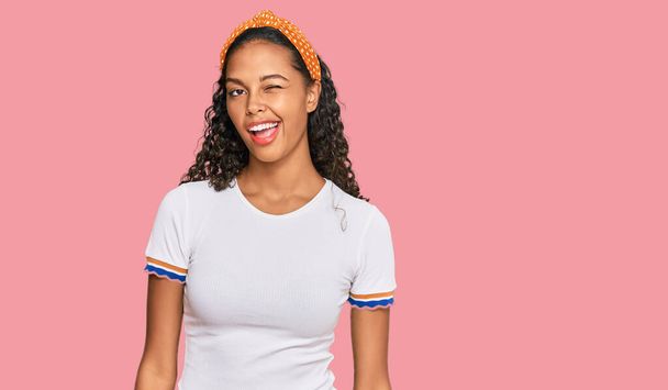 Νεαρή Αφροαμερικανίδα που φοράει καθημερινά ρούχα και κλείνει το μάτι κοιτάζοντας την κάμερα με σέξι έκφραση, χαρούμενο και χαρούμενο πρόσωπο.  - Φωτογραφία, εικόνα