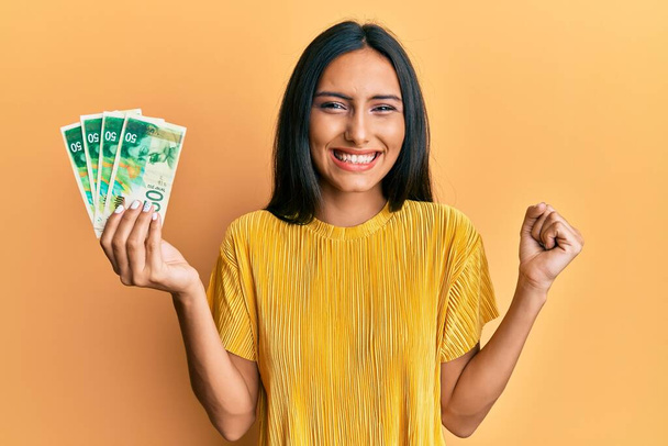 Junge brünette Frau mit 50 israel Schekel Banknoten schreit stolz, feiert Sieg und Erfolg sehr aufgeregt mit erhobenem Arm  - Foto, Bild