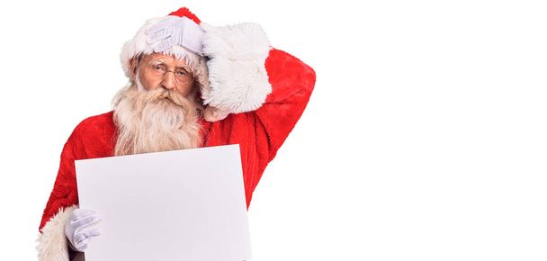 Viejo hombre mayor con pelo gris y barba larga con traje de Santa Claus sosteniendo pancarta estresado y frustrado con la mano en la cabeza, cara sorprendida y enojada  - Foto, Imagen