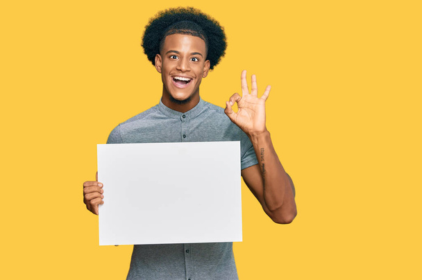 Африканский американец с африканскими волосами, держащий пустой баннер, делает хорошо знак с пальцами, улыбаясь дружелюбный жест отличный символ  - Фото, изображение