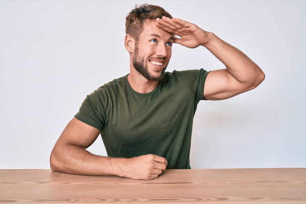 Young caucasian man wearing casual clothes sitting on the table very happy and smiling looking far away with hand over head. searching concept.  - Photo, Image