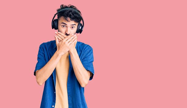 Молодий іспаномовний чоловік слухає музику, використовуючи навушники, шоковані, покриваючи рот руками за помилку. секретна концепція
.  - Фото, зображення